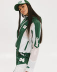A woman wearing a long-sleeved green and pink warm-up jacket with a hood, zipper pockets, and colorblocking. She also sports a matching green cap and appears to be standing against a white background.