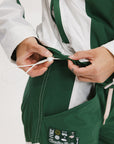 A woman wearing a long-sleeved green and pink warm-up jacket with a hood, zipper pockets, and colorblocking. She also sports a matching green cap and appears to be standing against a white background.
