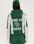 A woman wearing a long-sleeved green and pink warm-up jacket with a hood, zipper pockets, and colorblocking. She also sports a matching green cap and appears to be standing against a white background.