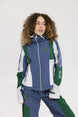 A female model wearing a blue and green color-blocked warm-up jacket with zipper pockets and a high neck collar.