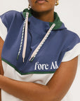 A person wearing a sleeveless hoodie with colorblocking and branding on the chest, in indigo, white, and green colors.