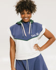 A person wearing a sleeveless hoodie with colorblocking and branding on the chest, in indigo, white, and green colors.