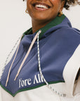 A person wearing a sleeveless hoodie with colorblocking and branding on the chest, in indigo, white, and green colors.