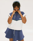 A person wearing a sleeveless hoodie with colorblocking and branding on the chest, in indigo, white, and green colors.