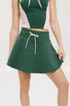A woman is wearing a green mini skirt with a round drawstring waist and silicone logos. She is also wearing a pink top and white sneakers.