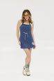 A woman is standing, wearing a sleeveless indigo dress with a round neckline, a drawstring at the waist, and white sneakers. She has glasses and her hair is styled.
