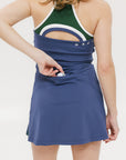 A woman is standing, wearing a sleeveless indigo dress with a round neckline, a drawstring at the waist, and white sneakers. She has glasses and her hair is styled.