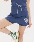 A woman is standing, wearing a sleeveless indigo dress with a round neckline, a drawstring at the waist, and white sneakers. She has glasses and her hair is styled.