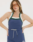 A woman is standing, wearing a sleeveless indigo dress with a round neckline and a drawstring at the waist.