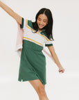 A woman in a green, white, and yellow striped polo dress with a ribbed collar and elastic toggles at the waist. The dress features side seam pockets and a branding stripe.