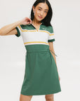 A woman in a green, white, and yellow striped polo dress with a ribbed collar and elastic toggles at the waist. The dress features side seam pockets and a branding stripe.