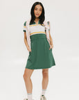 A woman in a green, white, and yellow striped polo dress with a ribbed collar and elastic toggles at the waist. The dress features side seam pockets and a branding stripe.