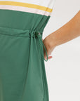 A woman in a green, white, and yellow striped polo dress with a ribbed collar and elastic toggles at the waist. The dress features side seam pockets and a branding stripe.