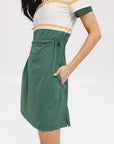 A woman in a green, white, and yellow striped polo dress with a ribbed collar and elastic toggles at the waist. The dress features side seam pockets and a branding stripe.