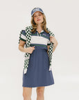 A woman wearing a polo dress with a color block design in white and indigo, featuring a ribbed collar, placket, and cuffs. She also wears a matching cap and sneakers.