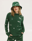 A woman wearing a green knit sweater with embroidered icons, matching green bucket hat, and green pants.