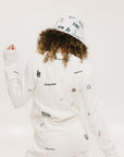 A woman wearing a white crew neck sweater with embroidered icons, matching white shorts, and a white bucket hat with a logo.