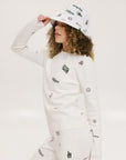 A woman wearing a white crew neck sweater with embroidered icons, matching white shorts, and a white bucket hat with a logo.