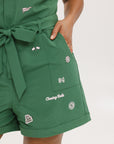 A woman wearing a green romper with embroidered golf icons, a tie belt at the waist, and cuffed sleeves.