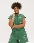 A woman wearing a green romper with embroidered golf icons, a tie belt at the waist, and cuffed sleeves. She is smiling and posing for the camera.