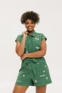 A woman wearing a green romper with embroidered golf icons, a tie belt at the waist, and cuffed sleeves. She is smiling and posing for the camera.