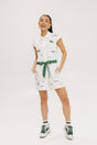 A woman is standing, wearing a white short-sleeved romper with embroidered golf icons, a tie belt at the waist, and white sneakers. She has dark hair and is smiling.