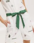 A woman is standing, wearing a white short-sleeved romper with embroidered golf icons, a tie belt at the waist, and white sneakers. 