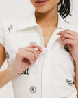 A woman is standing, wearing a white short-sleeved romper with embroidered golf icons, a tie belt at the waist, and white sneakers. 