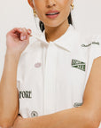 A woman is standing, wearing a white short-sleeved romper with embroidered golf icons, a tie belt at the waist, and white sneakers. 