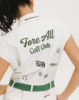 A woman is standing, wearing a white short-sleeved romper with embroidered golf icons, a tie belt at the waist, and white sneakers. 