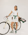 A woman is standing, wearing a white short-sleeved romper with embroidered golf icons, a tie belt at the waist, and white sneakers. 