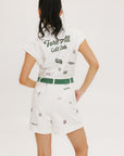 A woman posing in a white romper with embroidered golf-themed icons, a tie belt at the waist, and a zipper at the center front.