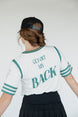 A person wearing a white golf shirt with green and black stripes, featuring a flamingo graphic and the text 'Fore All 04'. They are also wearing a black skirt and a black hat with text.