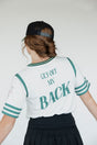 A person wearing a white golf shirt with green and black stripes, featuring a flamingo graphic and the text 'Fore All 04'. They are also wearing a black skirt and a black hat with text.