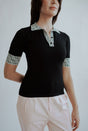 A woman wearing a black golf polo shirt with a geometric design on the collar and sleeves, and light pink pants. 
stylish women’s golf top outfit.